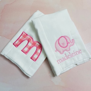 May include: Two white burp cloths with pink gingham embroidery. One cloth has the letter "M" and the other has a pink gingham elephant and the name "madeleine".