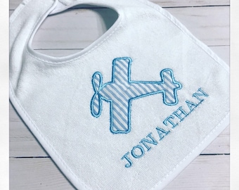 May include: White baby bib with a blue and white striped airplane design. The name "JONATHAN" is embroidered in blue below the airplane. The bib has a rounded shape with a white trim and a snap closure.