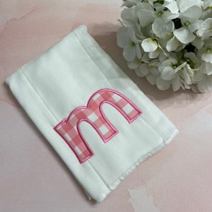 May include: White rectangular baby burp cloth with a pink gingham letter "M" applique. The letter is outlined in pink thread. The burp cloth is made of a soft, absorbent material.