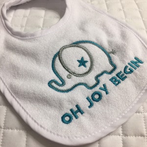 May include: White baby bib with a blue embroidered elephant and the text "OH JOY BEGIN".