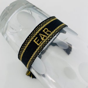 May include: Black and gold bracelet with the word "EAR" embroidered in gold. The bracelet is wrapped around a clear glass object. The bracelet has a gold tassel.
