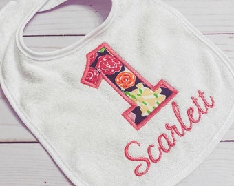 May include: White baby bib with a pink embroidered number 1 filled with a floral pattern. The name "Scarlett" is embroidered in pink script below the number. The bib has a hook and loop closure.
