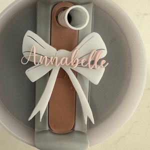 Puede incluir: Una tapa gris con un lazo blanco y el nombre "Annabelle" escrito en rosa. La tapa tiene un soporte de paja de plástico marrón.
