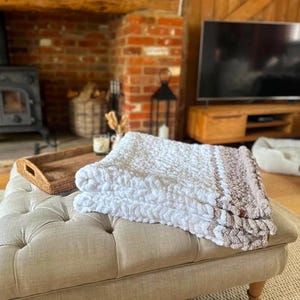 May include: A white and brown crocheted throw blanket with a textured pattern is folded on a light beige ottoman in a living room setting.