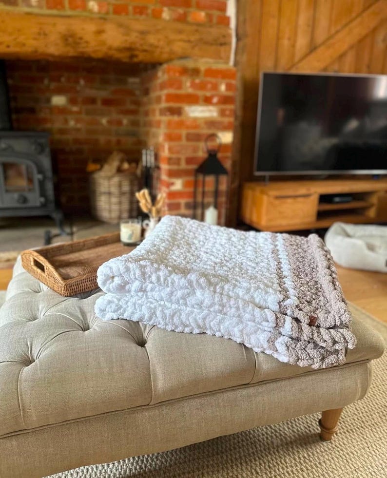 May include: A white and brown crocheted throw blanket with a textured pattern is folded on a light beige ottoman in a living room setting.