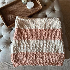May include: A pink and white striped chunky knit baby blanket. The blanket is on a light grey ottoman with a woven tray and candles on top.