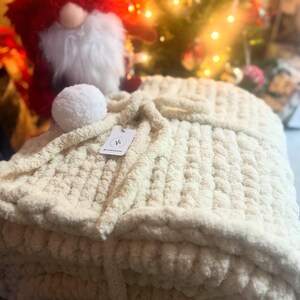 May include: A cream-coloured knitted throw blanket folded in a stack. The blanket has a fluffy texture and is tied with a cream-coloured ribbon. A small white tag with the letter "V" is attached to the blanket.