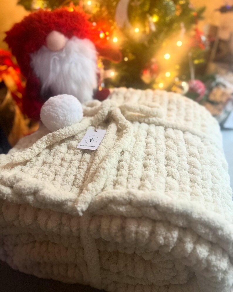 May include: A cream-coloured knitted throw blanket folded in a stack. The blanket has a fluffy texture and is tied with a cream-coloured ribbon. A small white tag with the letter "V" is attached to the blanket.