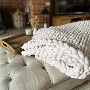 May include: A soft, white knitted throw blanket with a chunky texture, draped over a light grey tufted ottoman. The ottoman is in front of a brick fireplace and a television.