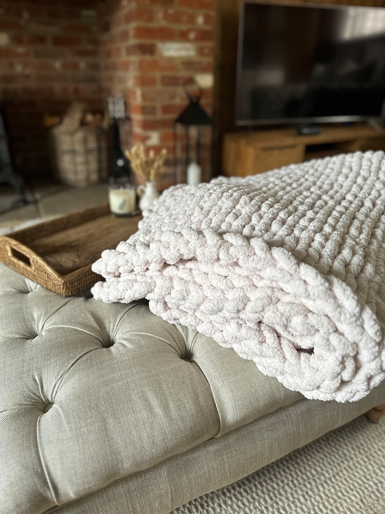 May include: A soft, white knitted throw blanket with a chunky texture, draped over a light grey tufted ottoman. The ottoman is in front of a brick fireplace and a television.