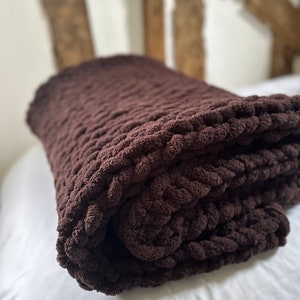 May include: A brown chunky knit throw blanket folded on a white bed. The blanket is made of thick yarn and has a soft, cosy texture.