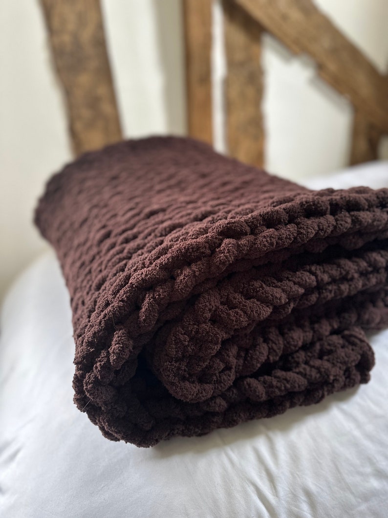May include: A brown chunky knit throw blanket folded on a white bed. The blanket is made of thick yarn and has a soft, cosy texture.