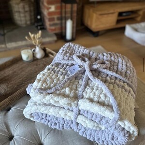 May include: A soft, chunky knit throw blanket in shades of white and light purple. The blanket is folded and tied with a ribbon.