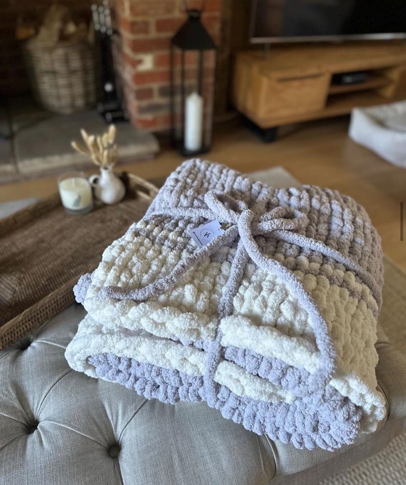 May include: A soft, chunky knit throw blanket in shades of white and light purple. The blanket is folded and tied with a ribbon.