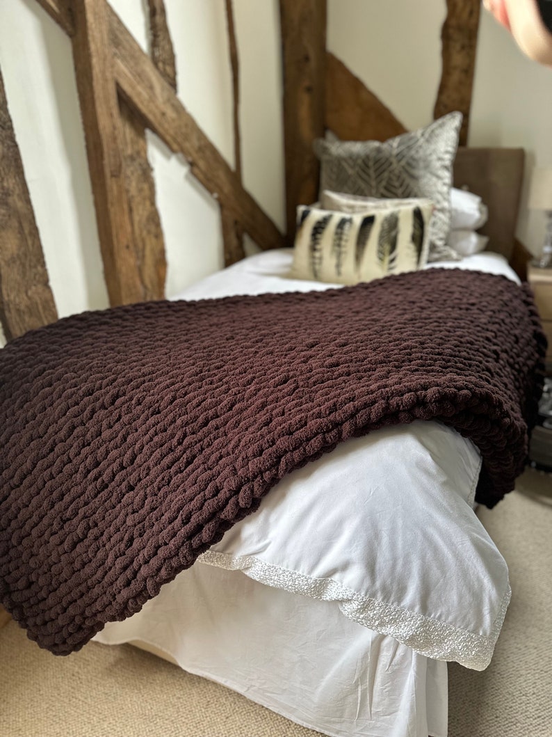 May include: A brown chunky knit throw blanket on a white bed with a white bed skirt. The bed has two pillows with a feather pattern on them.
