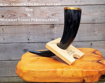 May include: A black Viking horn with a brass accent rim, resting on a personalized wooden stand. The stand is engraved with text. The horn is displayed on a wooden surface.