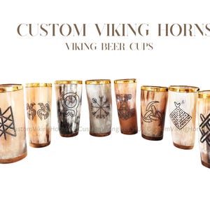 May include: A selection of Viking horn beer cups with gold rims and etched designs. The cups are made of natural horn material, with colours ranging from light beige to dark brown. The text "CUSTOM VIKING HORNS VIKING BEER CUPS" is displayed above the cups.
