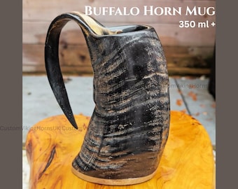 May include: A buffalo horn mug with a curved handle. The mug is black with brown and tan marbling. The text "BUFFALO HORN MUG 350 ml +" is at the top. The mug sits on a wooden surface.