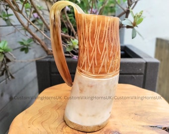 May include: A carved horn drinking mug with a light brown handle and a white body with a brown carved design. The mug is sitting on a wooden surface.