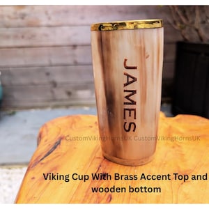May include: A Viking horn cup with a brass accent top and wooden base. The cup is engraved with the name "JAMES". The cup is made of a light brown material and sits on a wooden surface. The cup is approximately 15 cm tall.