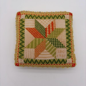 May include: A square needlepoint pincushion featuring a geometric star pattern in shades of orange, olive green, and cream. The border displays a zigzag stitch in green and red, with the embroidered text JBC 1981 visible in the bottom right corner.