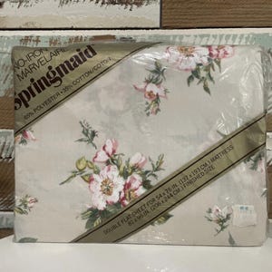 Pode incluir: Um lençol plano duplo Springmaid embalado. O lençol apresenta um padrão floral com flores rosa e brancas sobre um fundo bege claro. A embalagem está rotulada "No-Iron Marvelaire" e é para um colchão de 137 x 193 cm.