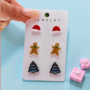 May include: A set of Christmas-themed stud earrings on a white card. The earrings include Santa hats, gingerbread men, and Christmas trees. The word "JEWELRY" is printed at the top of the card. Two small pink gift boxes with white ribbons are also present.