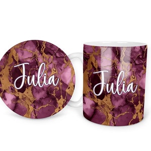 May include: A white ceramic mug and coaster with a pink and gold marble design. The mug and coaster both have the name "Julia" written in white cursive.