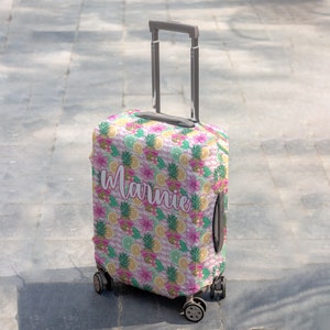 May include: A pink, green and yellow luggage cover with a pineapple and citrus fruit pattern. The cover has the name "Marnie" printed on it.