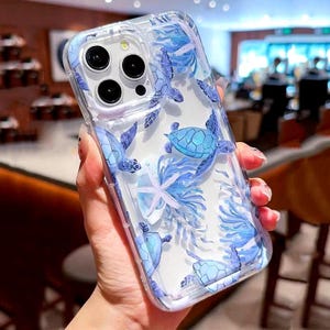 Puede incluir: Una funda transparente para teléfono con un diseño azul y blanco con tortugas marinas y estrellas de mar. La funda está diseñada para un teléfono con una cámara de triple lente.