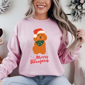 May include: A pale pink sweatshirt with a cartoon dog wearing a Santa hat and a green bandana. The text "Merry Woofmas" is written in red and pink. This festive sweatshirt is part of a holiday clothing range.