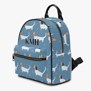 May include: A blue backpack with a repeating pattern of dachshund dogs in various poses. The backpack features black trim, a black handle, and a zipper closure. The initials "KMH" are displayed on the front of the bag.
