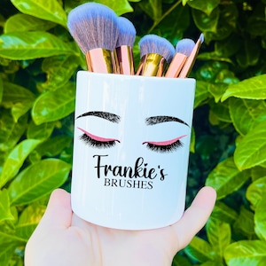 Puede incluir: Portacepillos de maquillaje de cerámica blanca con un diseño de ojo negro y rosa y el texto "Frankie's Brushes".
