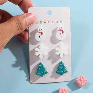 May include: A set of Christmas-themed earrings on a white card. The earrings include snowman, snowflake, and Christmas tree designs. Two small pink gift boxes are also visible. The word "JEWELRY" is printed at the top of the card.