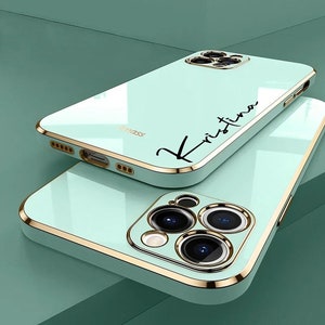 Op de afbeelding: Een mintgroene telefoonhoes met een gouden rand en een zwarte inscriptie die "Kristina" zegt. De hoes heeft een verhoogde camerabult met drie lenzen.