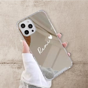 Può includere: Una custodia per telefono trasparente con finitura a specchio. La custodia ha un cuore bianco e il nome "Remi" scritto su di essa.