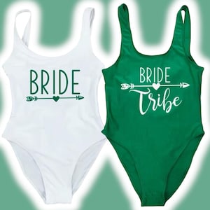 May include: Two white one-piece swimsuits with green text. One swimsuit says "Bride" with an arrow and heart. The other swimsuit says "Bride Tribe" with an arrow and heart.