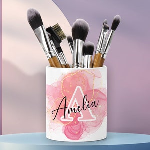 Puede incluir: Portacepillos de maquillaje de cerámica blanca con un diseño de acuarela rosa y dorado. El portacepillos presenta la letra "A" y el nombre "Amelia" en escritura negra. El portacepillos está lleno de varios pinceles de maquillaje.