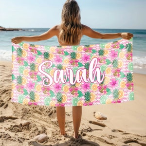 May include: A pink beach towel with a tropical print of pineapples, oranges, and flowers. The name "Sarah" is written in white cursive on the towel.