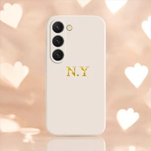 Puede incluir: Funda de teléfono blanca con un diseño N.Y. dorado y una cámara de triple lente.