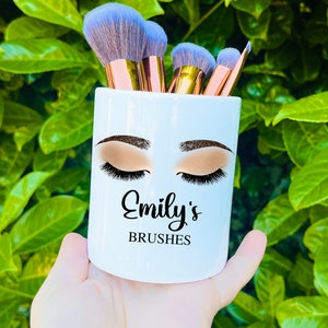 May include: White ceramic makeup brush holder with a stylized illustration of a woman's eyes with eyelashes and the text "Emily's Brushes".