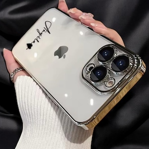 May include: A clear phone case with a gold trim and a black camera lens cover. The case has a black spade and the word "Justine" written on it.