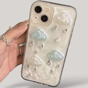 以下が含まれることがあります： 真珠光沢のある白い背景に、水色の雲と透明な涙型の装飾が施された透明な携帯電話ケース。ケースは透明な縁取りがされています。