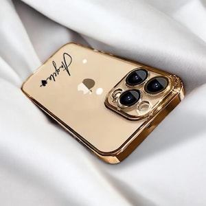May include: A gold iPhone case with a clear back and a black spade and the word "Apple" written in cursive. The case has a gold trim around the edges and a gold camera ring.
