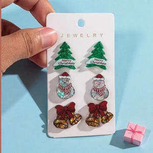 May include: A set of Christmas-themed stud earrings on a white card. The earrings feature green Christmas trees with "Merry Christmas" text, snowmen, and gold bells with red bows. A small pink gift box is also present.
