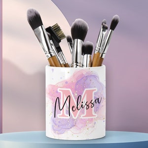 May include: White ceramic makeup brush holder with a pink and purple watercolor design and gold glitter. The holder has the name "Melissa" written on it with a pink "M".