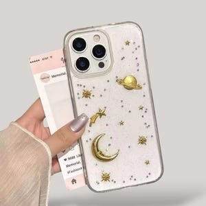 May include: Clear phone case with a white glitter background and gold celestial accents, including a planet, stars, and a crescent moon. The case has a clear border and a camera cutout.