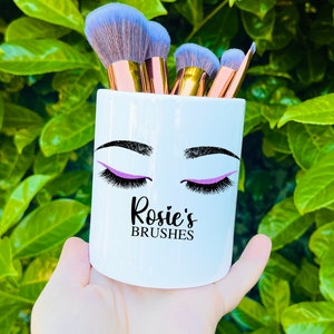 May include: White ceramic makeup brush holder with a black and purple eye design and the text "Rosie's Brushes".
