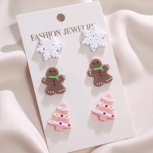 May include: A set of Christmas-themed stud earrings on a white card. The earrings include white snowflake shapes, gingerbread men with green scarves, and pink Christmas trees with colorful ornaments. The card reads "FASHION JEWELRY".