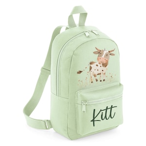 May include: A light green canvas backpack with a brown and white cow illustration and the name "Kitt" printed on the front pocket.